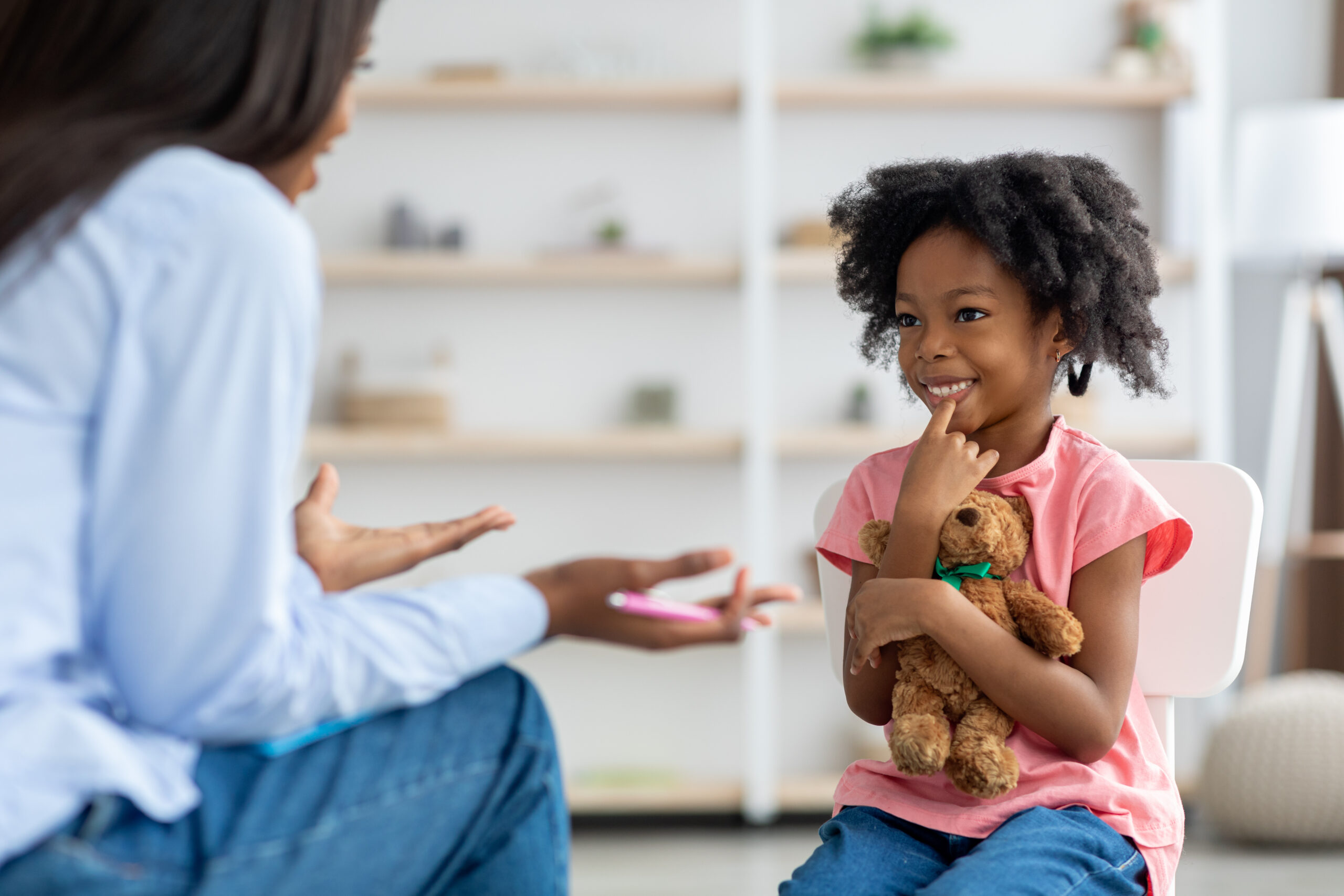 Cheerful cute little african american girl at child psychologists office, sitting on chair in front of unrecognizable female therapist, hugging teddy bear, smiling, feeling safe. Child psychotherapy